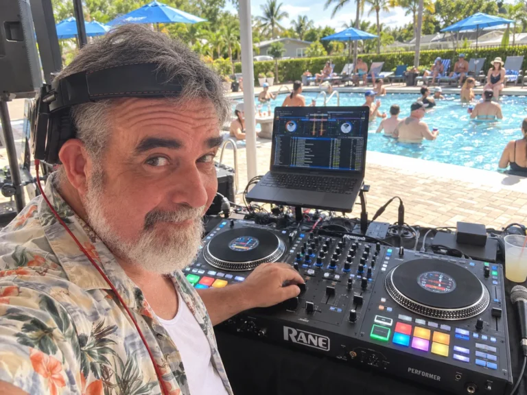 DJ 2RO at the DJ booth during a daytime pool party with blue umbrellas